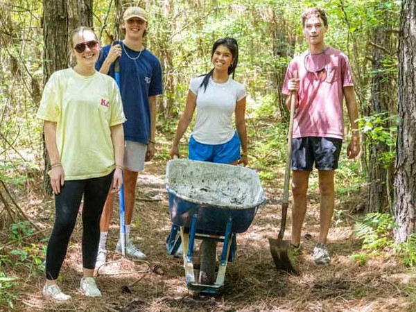 A group of students walks along a woodland path carrying shovels and pushing a wheelbarrow.