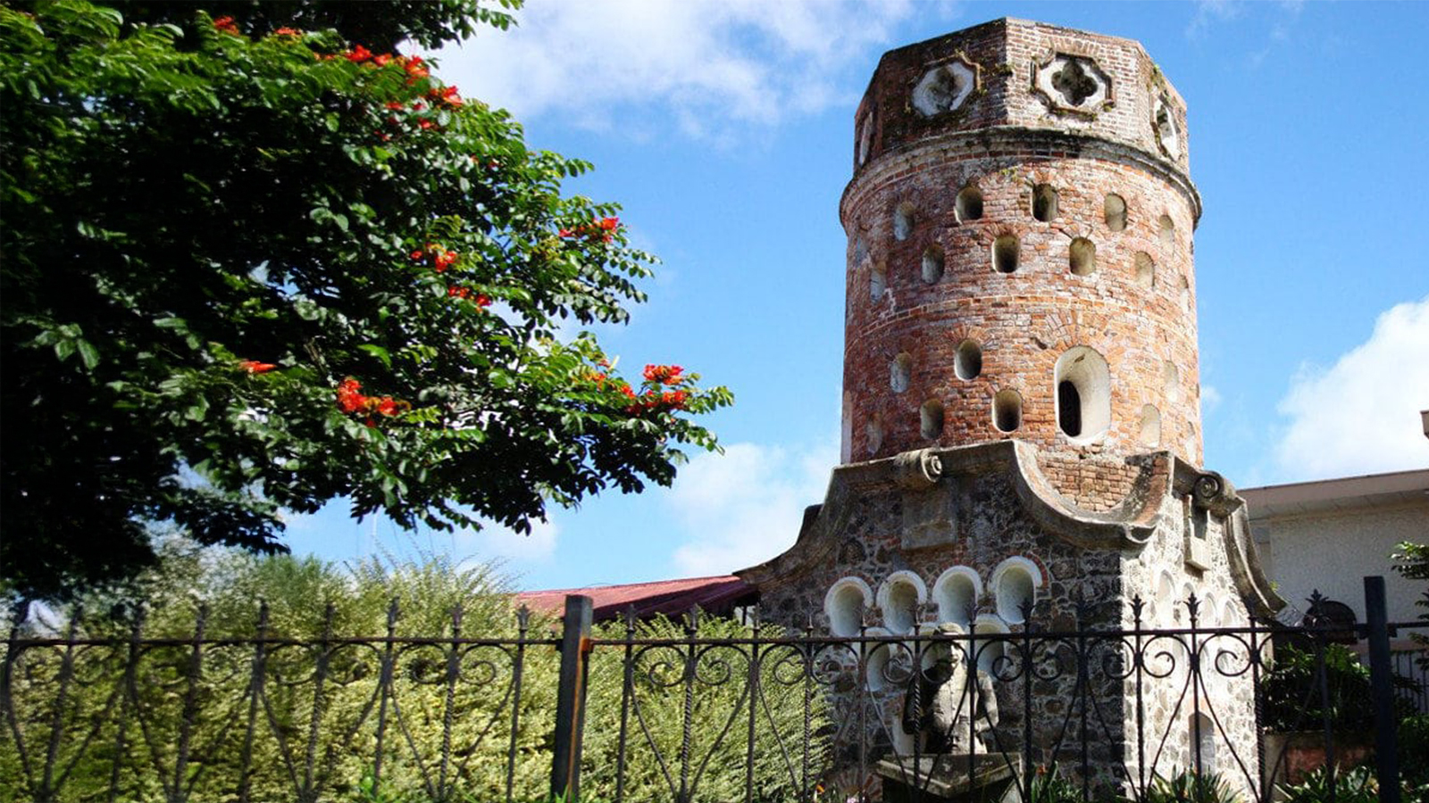 Historic brick tower in Costa Rica surrounded by greenery and an iron fence under a bright blue sky.