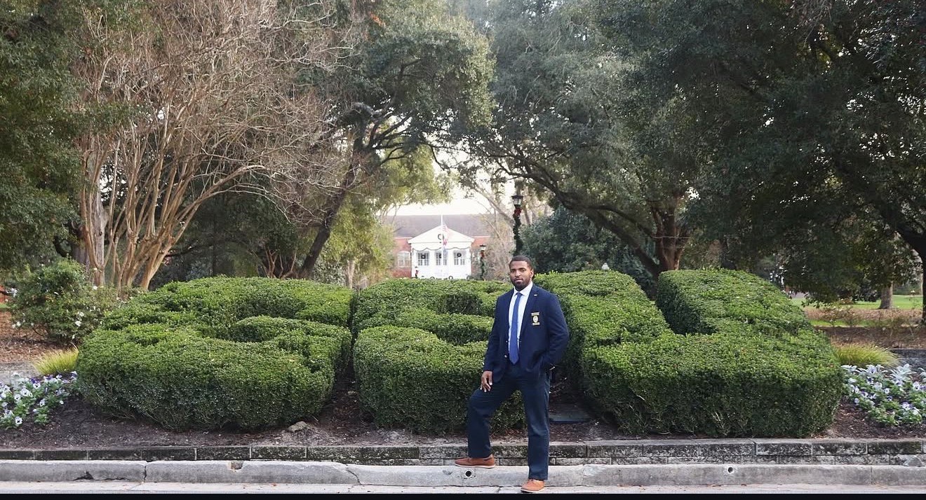 University Police Investigator Heiko Ellis stands in front of the GSU bushes on Sweetheart Circle at Georgia Southern University. He is wearing a dark blue suit and his badge.