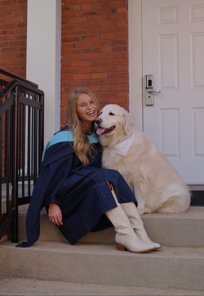 Madison Buford smiles while sitting on a staircase with her golden retriever, Sophie. Madison is wearing her graduate regalia for her upcoming commencement ceremony, where she will be awarded her master's degree.