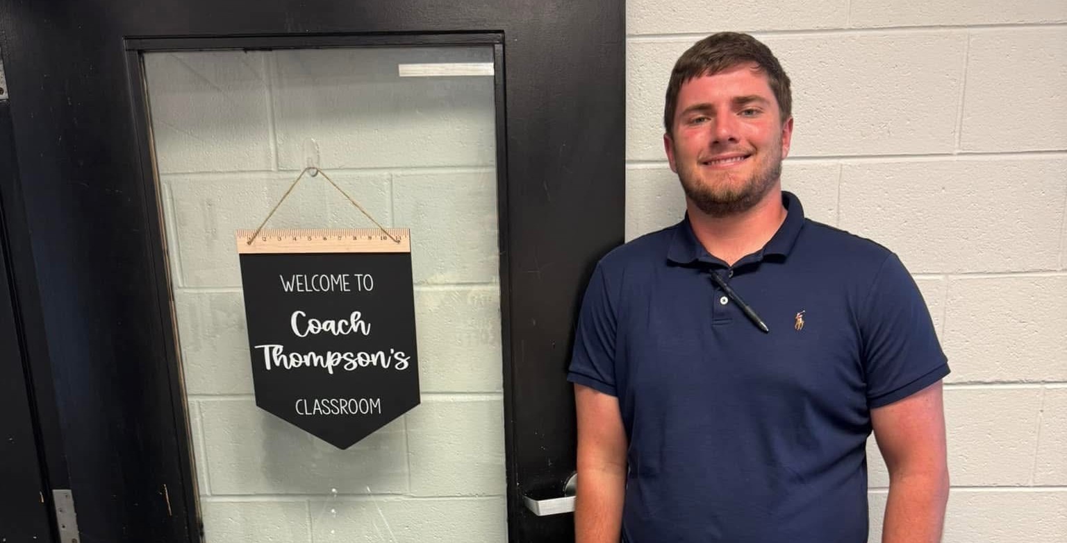 Man in blue shirt and khaki pants poses next to his classroom door with the name "Coach Thompson" on it
