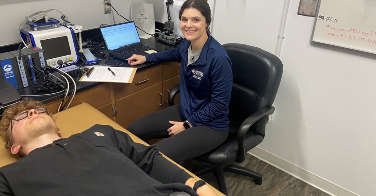 Georgia Southern graduate student Allie Wade sits at a computer monitoring a device worn on the wrist of a subject laying flat on a table in front of her