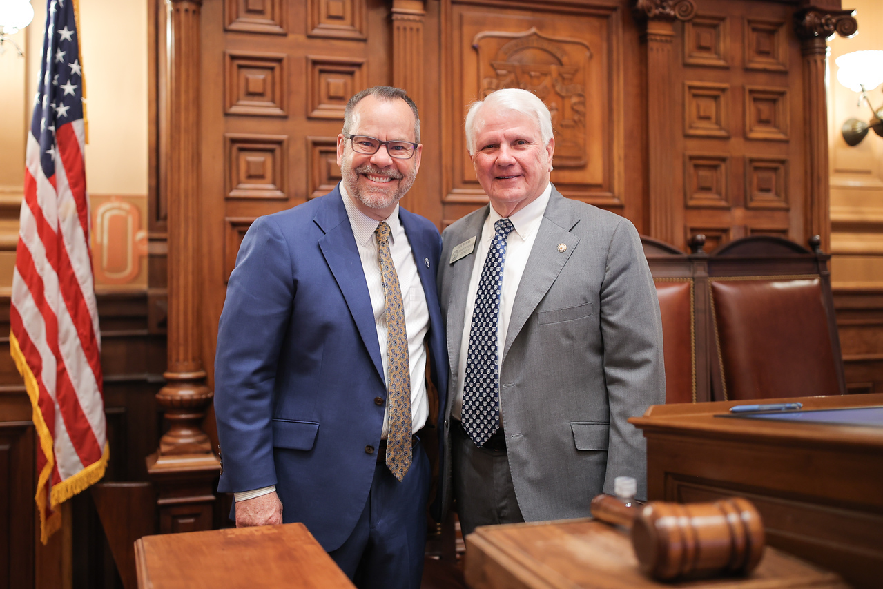 Two men in suits smile and pose together in a wood-paneled government chamber with an American flag in the background