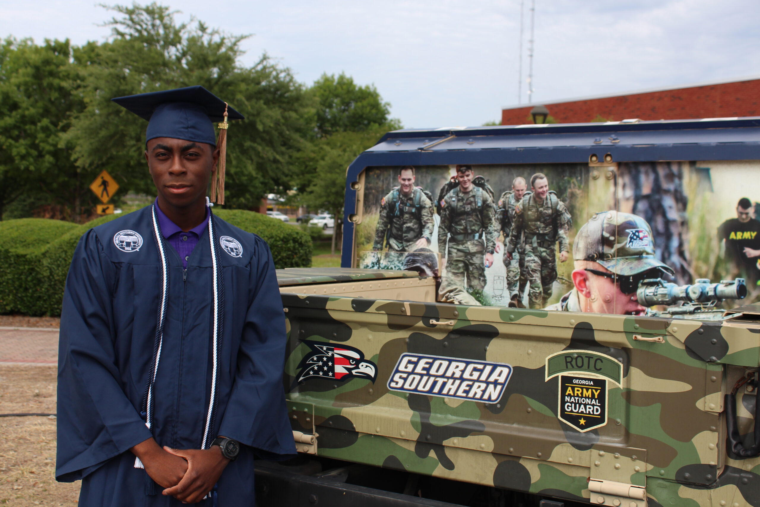 A graduate in all blue graduation regalia and a gold tassel with a gold cord stands next to a camo military truck with the georgia southern logo proudly