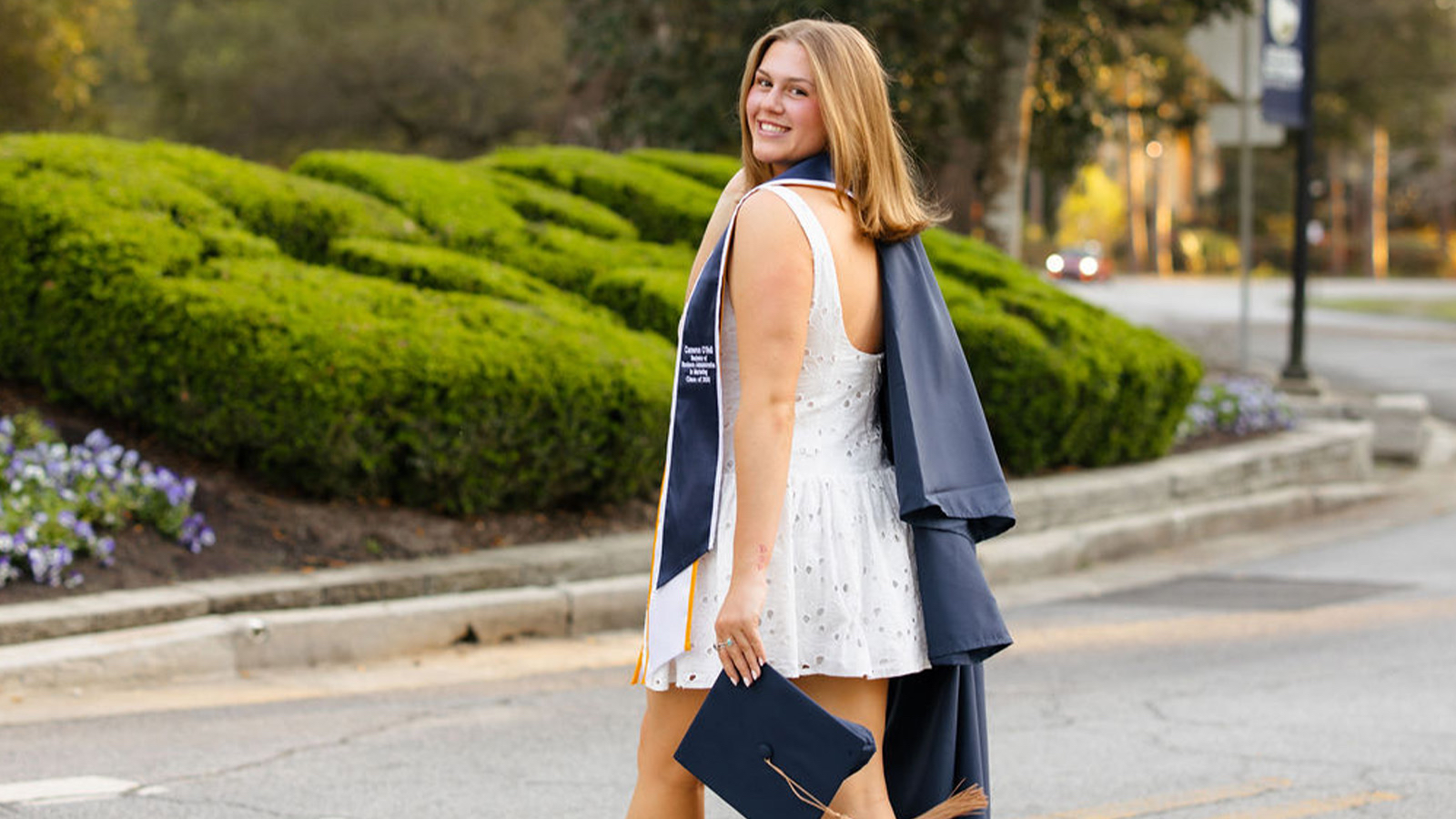 Smiling graduate in a white dress with a navy robe over her shoulders walks along a street, holding a graduation cap and gown accessories.