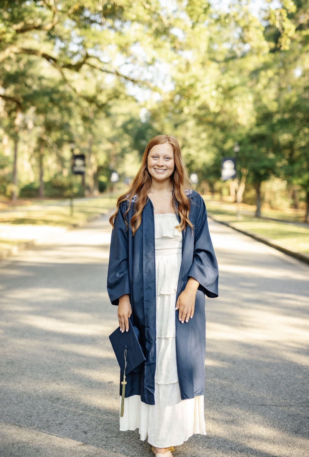 Smiling young woman in a navy graduation gown standing on a tree-lined path with a cap in hand, celebrating outdoors.