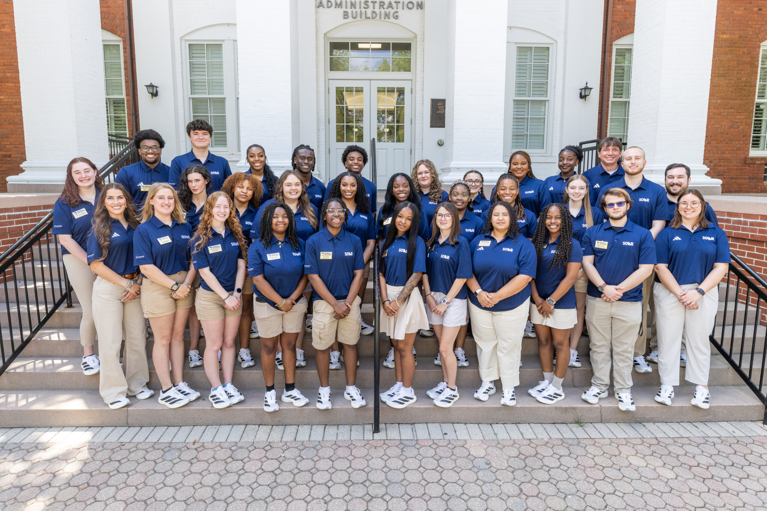 Group of students in blue polo shirts posing on the steps of a white brick administration building, smiling at the camera.