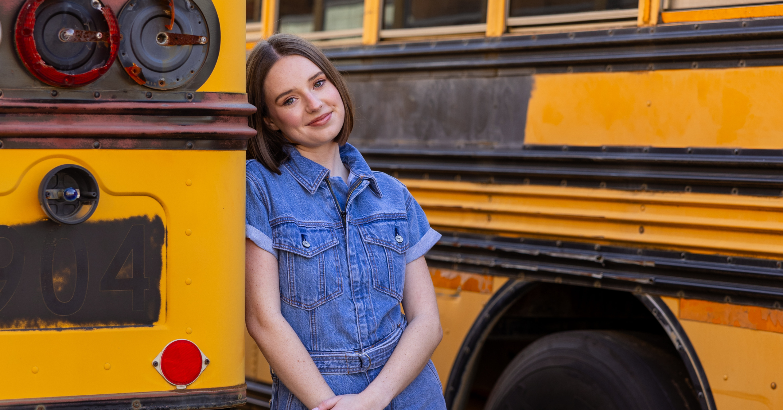 Georgia Southern student Ashley Claas leans against an old yellow school bus. She has short brown hair and is wearing a denim outfit while smiling kindly at the camera.