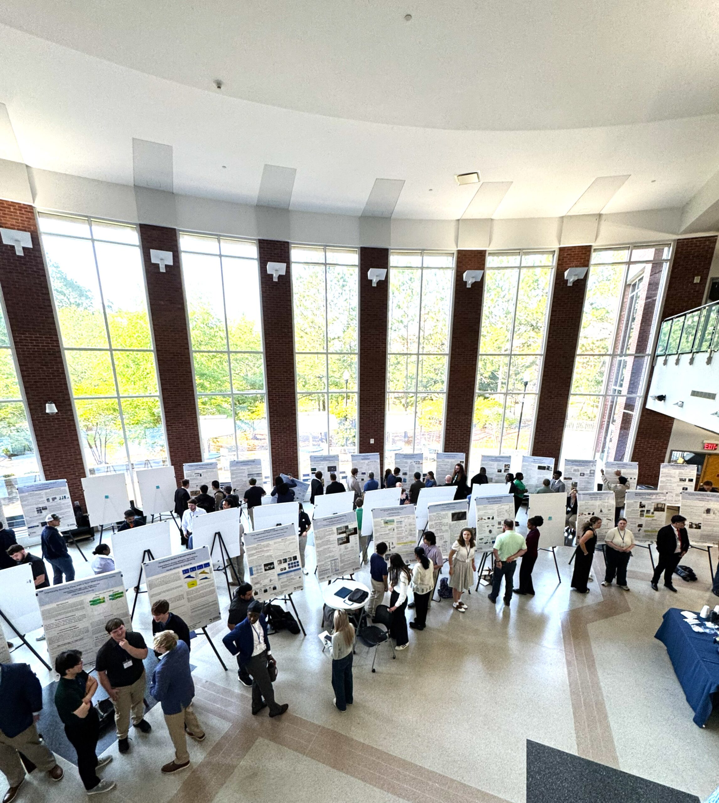 Students in the College of Engineering atrium standing beside research posters. The atrium has large windows shpwing trees.