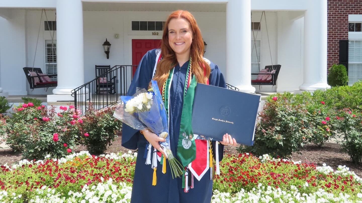Jaylee Kilgo stands in front of her sorority house while wearing commencement regalia and holding flowers and a diploma cover.