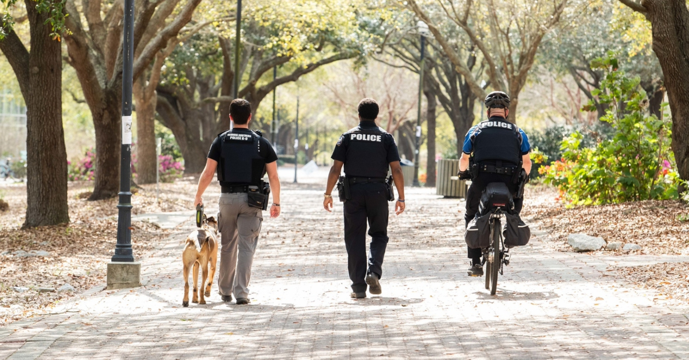 Three police officers walk along a tree-lined park path; a police dog walks with the left officer while a cyclist in police gear follows on the right with a bike helmet on.