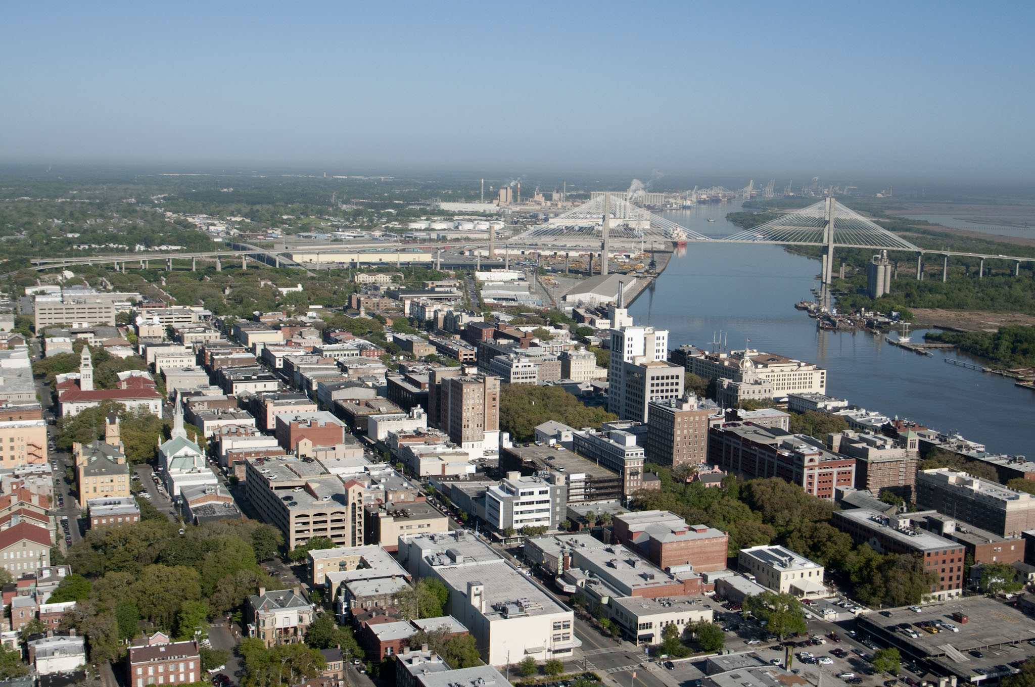 Aerial view of a city with a wide river and a cable-stayed bridge spanning it toward industrial port facilities in the distance.