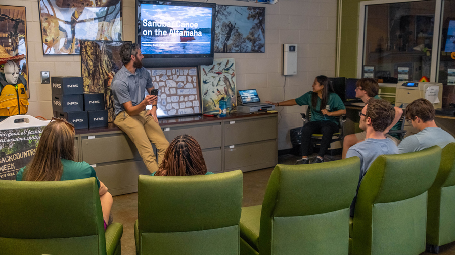 Presenter sits on a cabinet with a tablet, leading a slide presentation to a small group in a classroom-like room with green chairs.