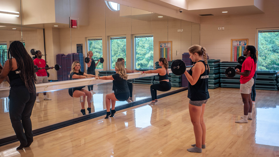 Group fitness class in a mirrored gym; participants hold a long bar across their shoulders and squat while an instructor supervises nearby.