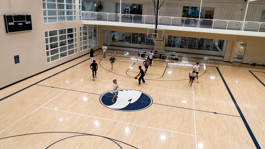 Indoor basketball gym viewed from above with players on the court, a large blue-and-white logo at center, and a balcony running along the second floor.