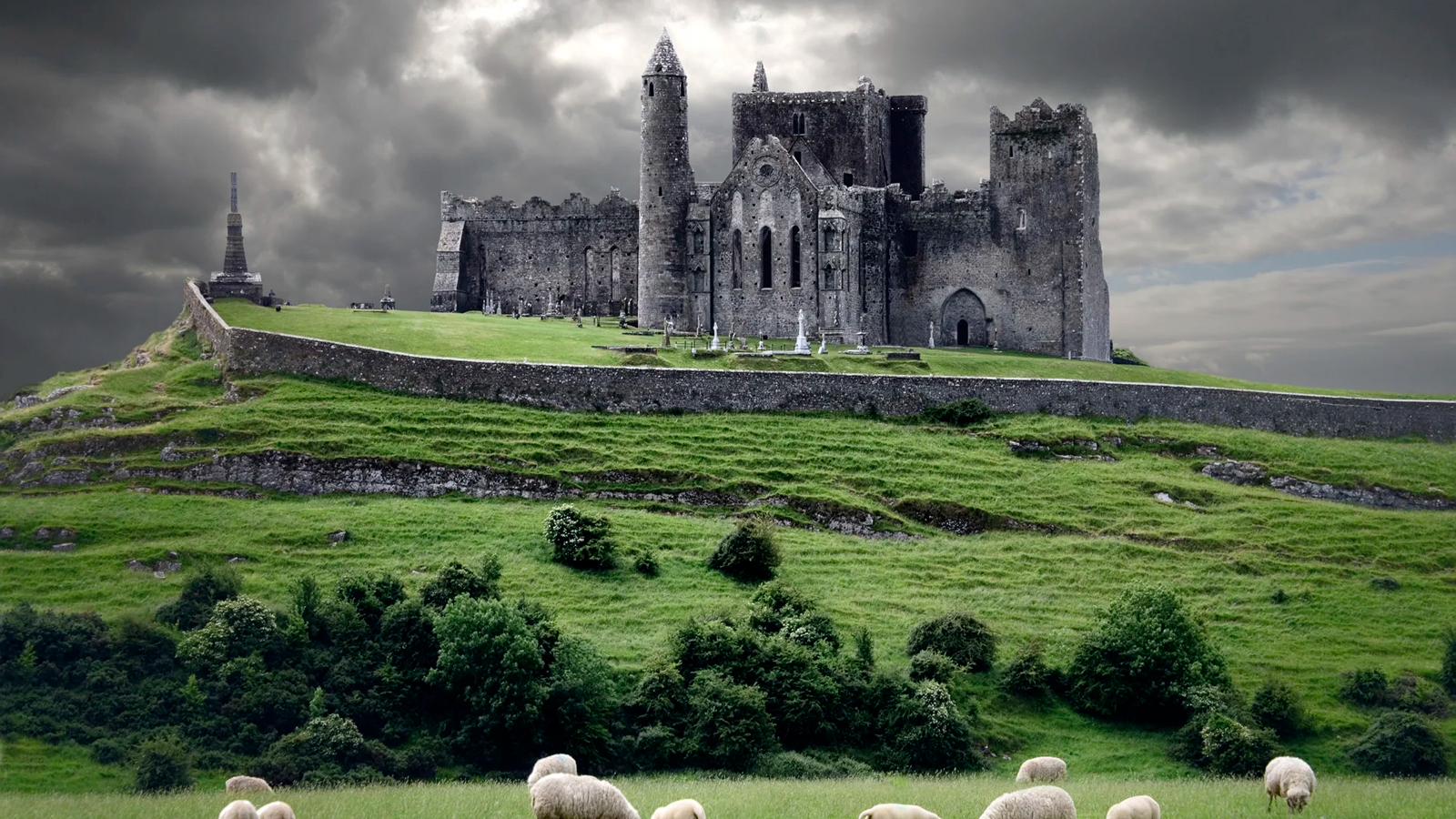 Ruined medieval castle atop a grassy hill with sheep in the foreground under dark storm clouds.