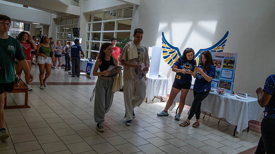 People walk through a bright indoor hallway of a campus event with tables and posters along the walls.