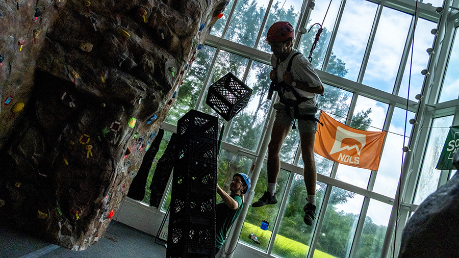 Climber in harness and helmet suspended mid-air indoors while another person belays nearby, with a large textured climbing wall and orange banner in the background.