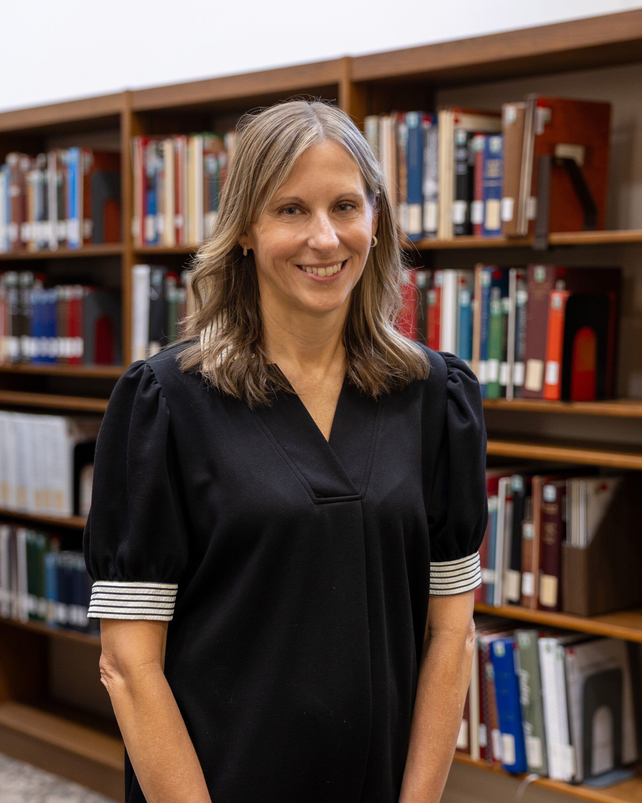 Smiling woman in a black top stands in a library with shelves of books behind her.