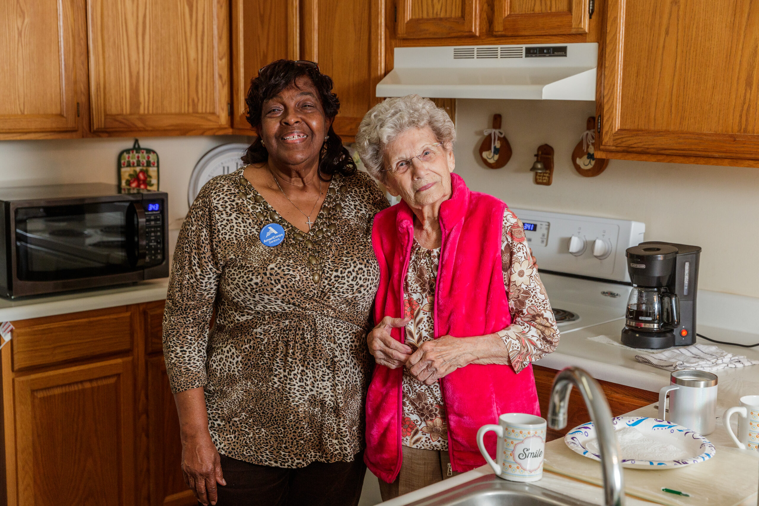 Two women stand together in a warm kitchen, smiling at the camera; one wears leopard print, the other a bright pink vest.