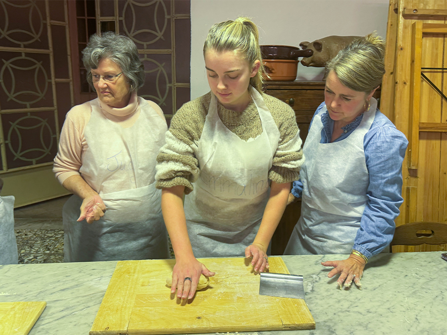 Three women in aprons stand at a marble counter, cutting dough on wooden boards during a cooking class.