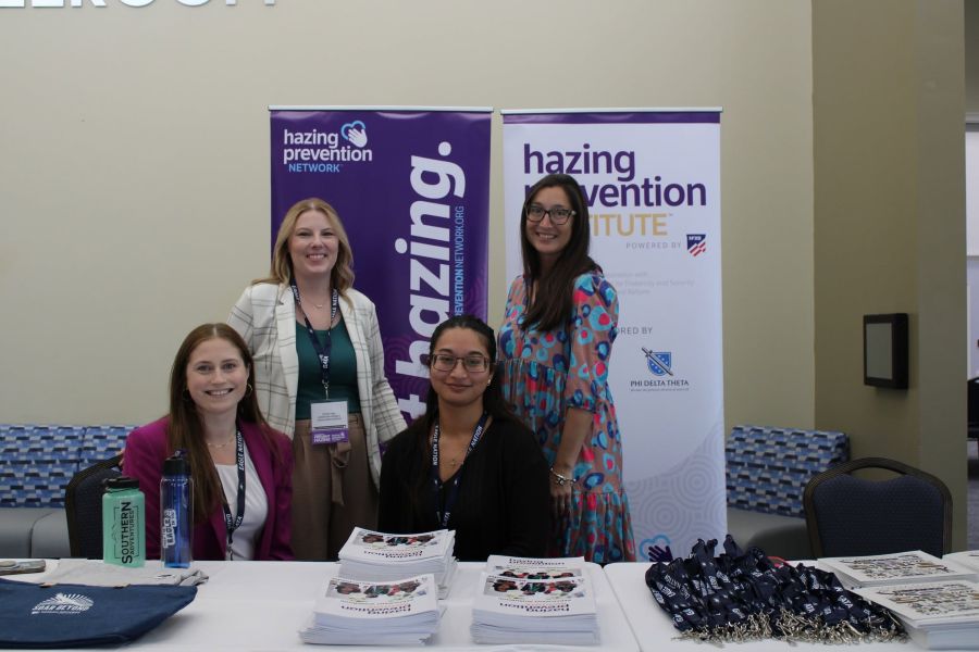 Four women at a hazing-prevention information booth, standing and smiling in front of banners and tables of pamphlets and swag.