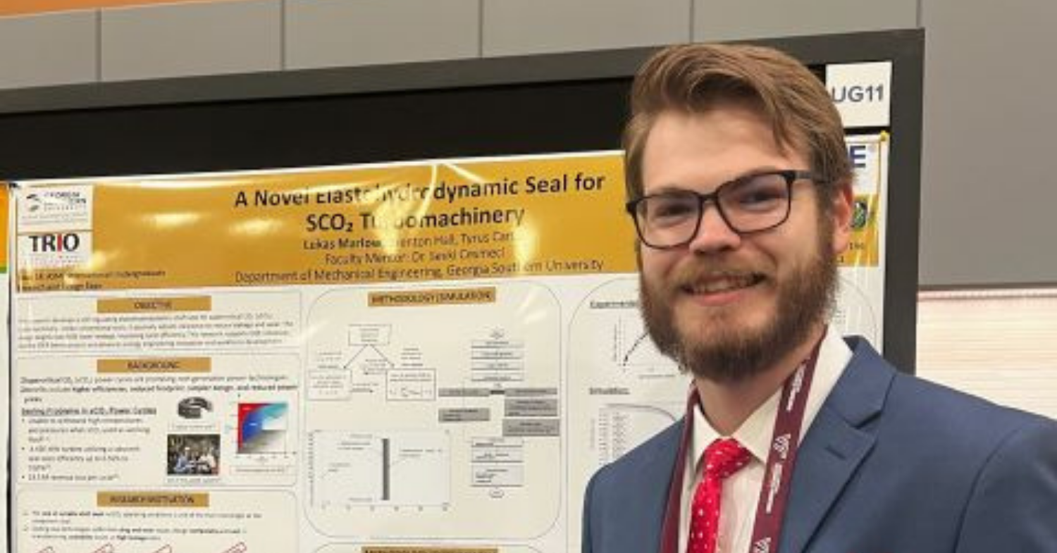 Man with glasses and a beard in a blue suit and red tie stands beside an academic poster at a conference. This is an informative image of a presenter with a poster.