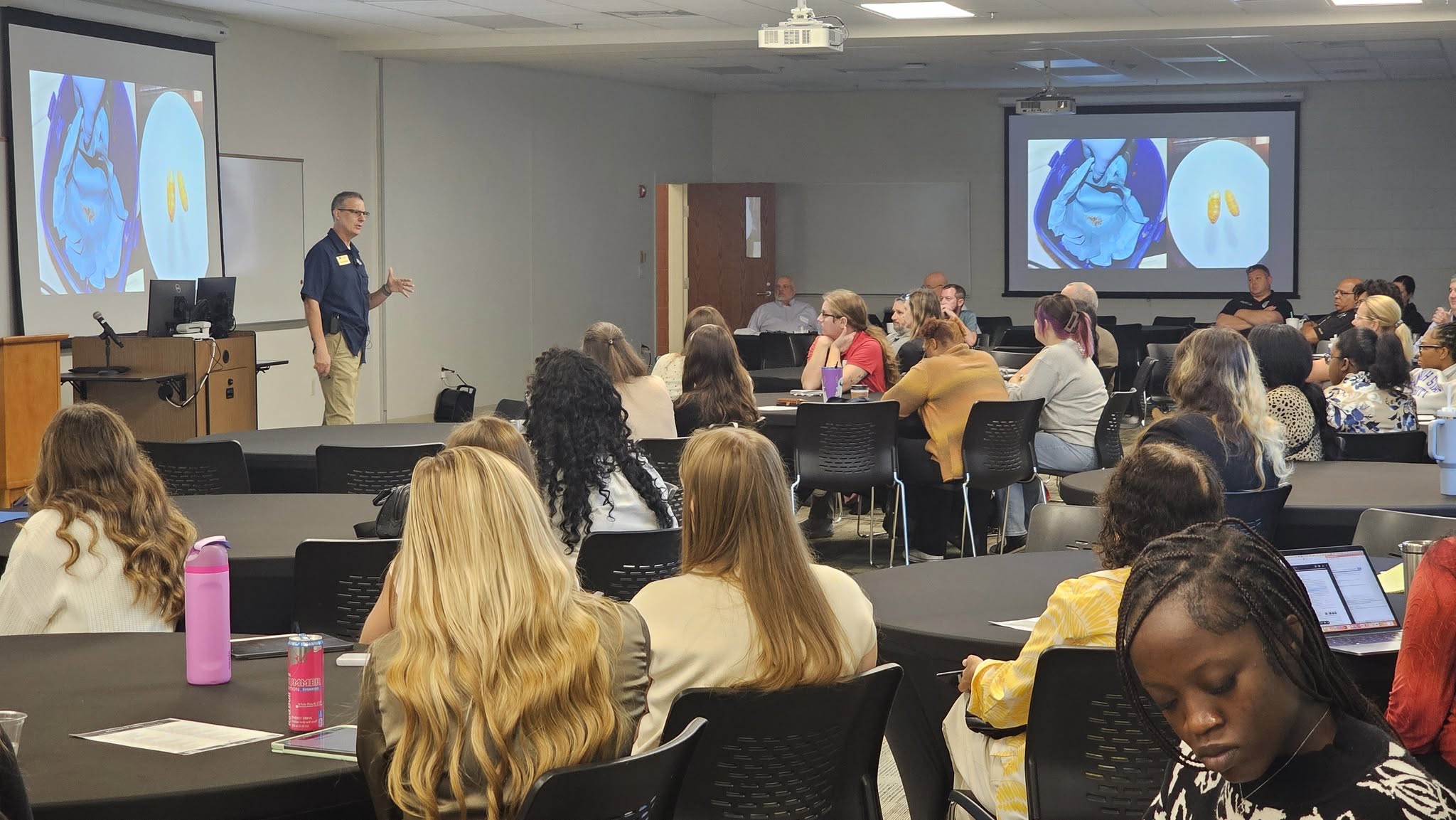 Professor Ed Mondor stands in front of a large room. An audience of a few dozen people are seated at tables around the room. A projector screen shows an image of a medical scan.