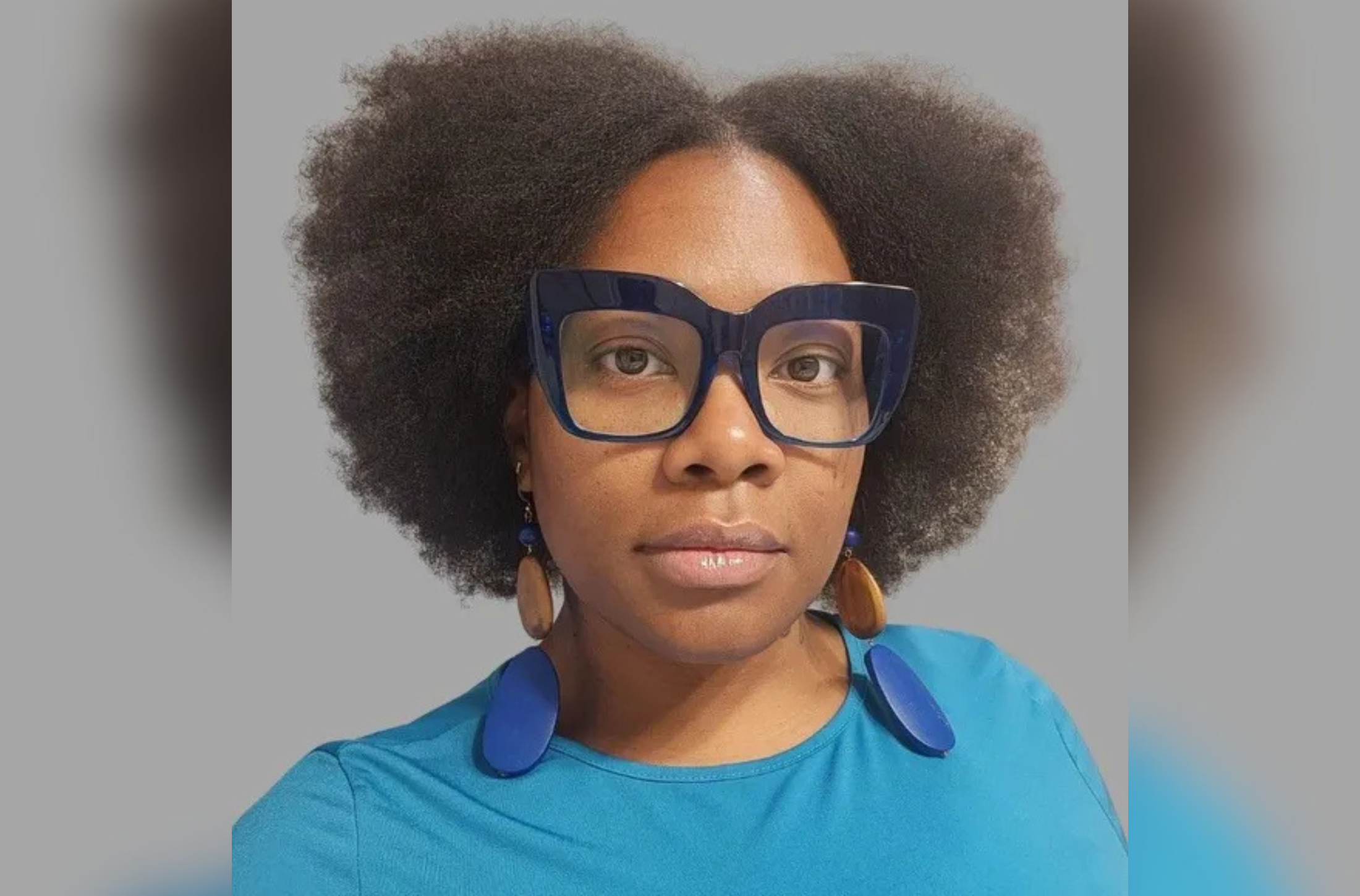 The photo features a portrait of a woman with beautifully shaped natural hair framing her face. She is wearing dark blue, thick-rimmed glasses which compliments her ocean blue top and wide, round-shaped dangly earrings which alternate between dark blue and light brown oval shapes.